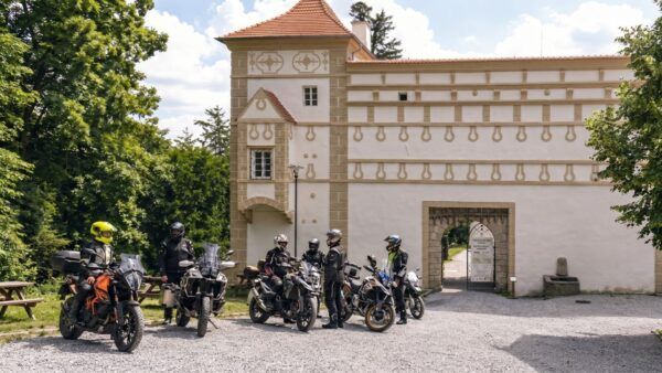 Skupina motorkářů se zaparkovanými stroji před historickou budovou EFI Zámku Račice. Zámek nabízí bezpečné parkování a stylové ubytování pro motorkáře na jižní Moravě.