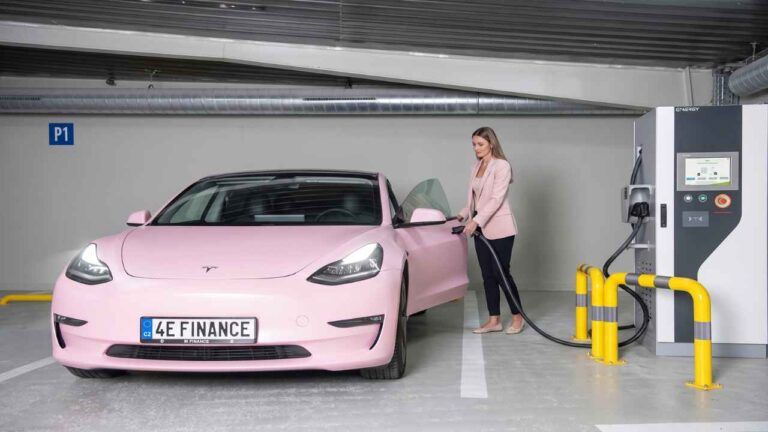 Růžový elektromobil u nabíjecí stanice v garáži a žena vedle vozu.