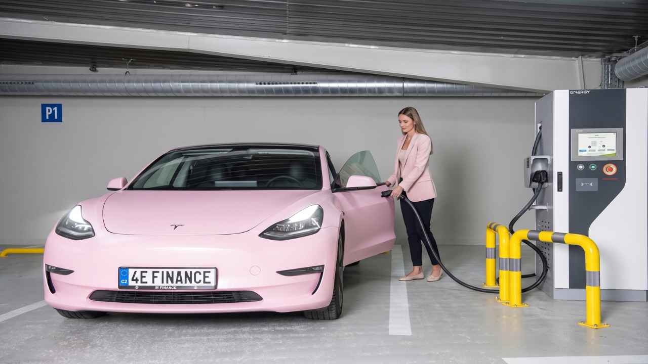Růžový elektromobil u nabíjecí stanice v garáži a žena vedle vozu.